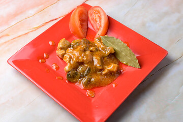 Pork stew with a bay leaf and tomato garnish on a red plate, marble background, typical food, typical mediterranean mallorcan cuisine typical from balearic islands mallorca, spain