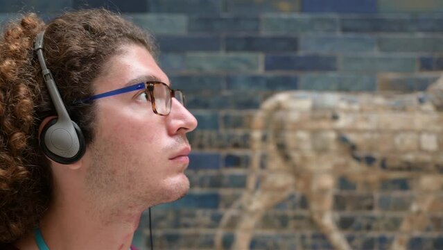 Berlin, Germany, August 16, 2023. Portrait of a young caucasian man with curly hair and glasses busy listening to the audio guide with headphones at the Ishtar Gate in the Pergamon Museum in Berlin.