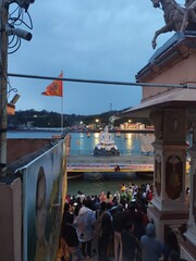La Maha Kumbh Mela, réunion et célébration religieuse, près du Gange, la nuit, le soir, lumière allumés, sous un temps pluvieux, respect des dieux, ciel nuagueux, avec tour indiquant les heures, décor