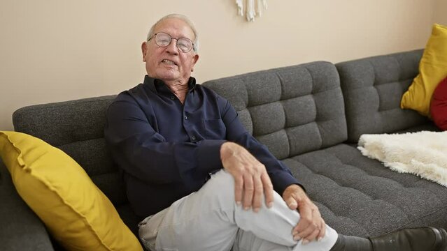 Cheerful senior man resting at home, confidently speaking and expressing joy, comfortably sitting on living room sofa, enjoying a relaxed indoor conversation