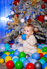 Fototapeta na wymiar Small child sits in ball pit near the Christmas tree and holds Christmas ball in his hands.