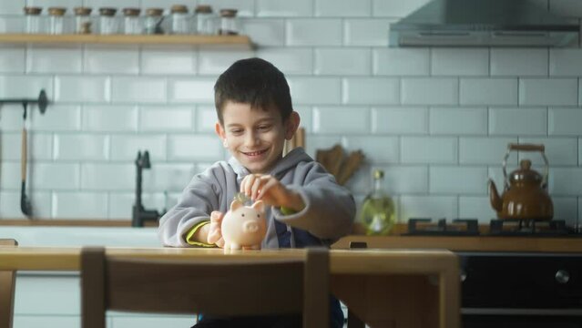 Smiling boy sit in the kitchen putting coins into a piggy bank. The child saves money and invests in the future. Concept of teaching financial literacy, budget planning