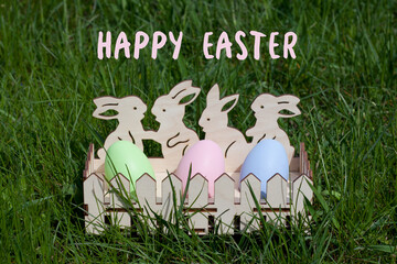 Greeting card with text Happy Easter in English. Cute wooden egg cup with painted Easter eggs standing on green grass in spring garden. Easter tradition of egg hunting. Outdoor