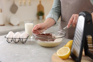 Woman making chocolate dough with whisk in bowl at gray marble table, closeup