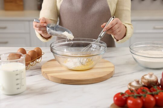 Woman whisking eggs while adding flour into bowl at light marble table indoors, closeup