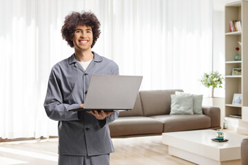 Fototapeta premium Young man in pajamas standing in a living room with a laptop computer