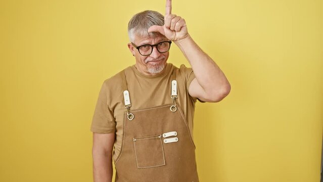Grey-haired, middle-age man in glasses and apron mockingly insults people with loser gesture, forehead fingers standing against isolated yellow background