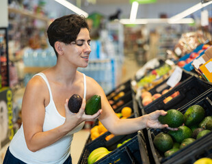 Positive interested young Latina choosing ripe avocado from plastic boxes on supermarket showcase ..