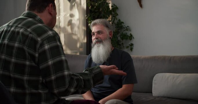 A happy man with gray hair and a lush gray beard has fun and rejoices during his communication with his boyfriend a brunette with stubble in a plaid shirt sitting on a gray sofa in a modern apartment