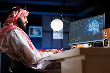 Islamic man uses AI and machine learning to ensure cyber security for a network. He monitors data centers and servers working with a PC monitor displaying advanced software and a neural network.