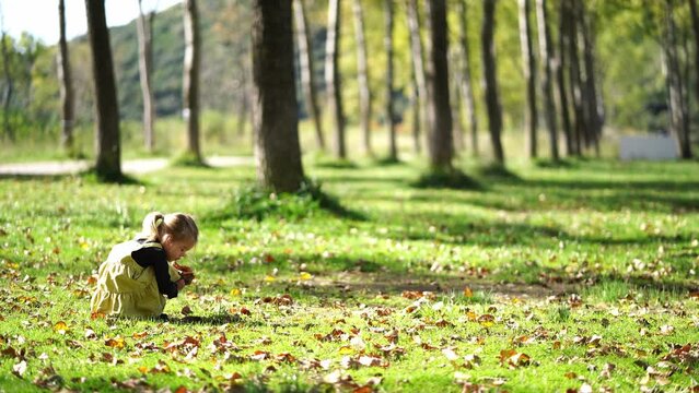 A happy little girl is sitting on the green grass in a natural landscape park, surrounded by trees and plants, enjoying the peaceful atmosphere