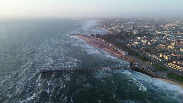 Aerial drone film of the town Swakopmund in Namibia, Africa during sunset. Massive dramatic ocean rolling waves crashing on the sand beach. Long wooden pier, desert and red sand dunes