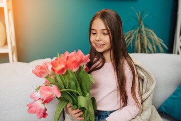 Little cute girl holding bouquet of pink tulips while sitting on sofa at home
