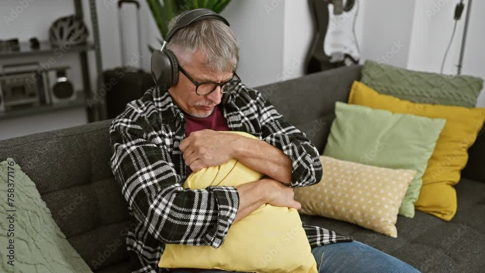 Sticker A mature man in glasses and headphones embraces a yellow pillow on a sofa at home, signifying solitude or relaxation.