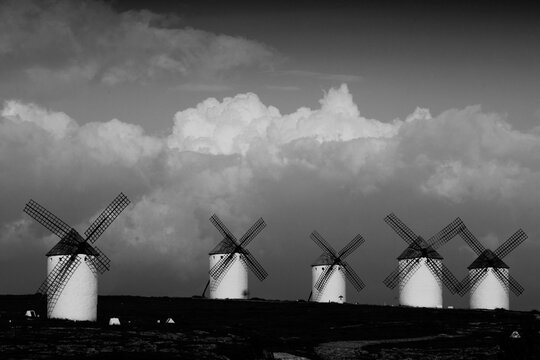 Traditional windmills lined up on a plain, their sails standing out against fluffy clouds, captured in stunning black and white