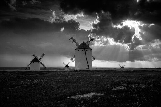 Monochrome landscape featuring a row of traditional windmills under a dramatic sky with sunbeams piercing through clouds