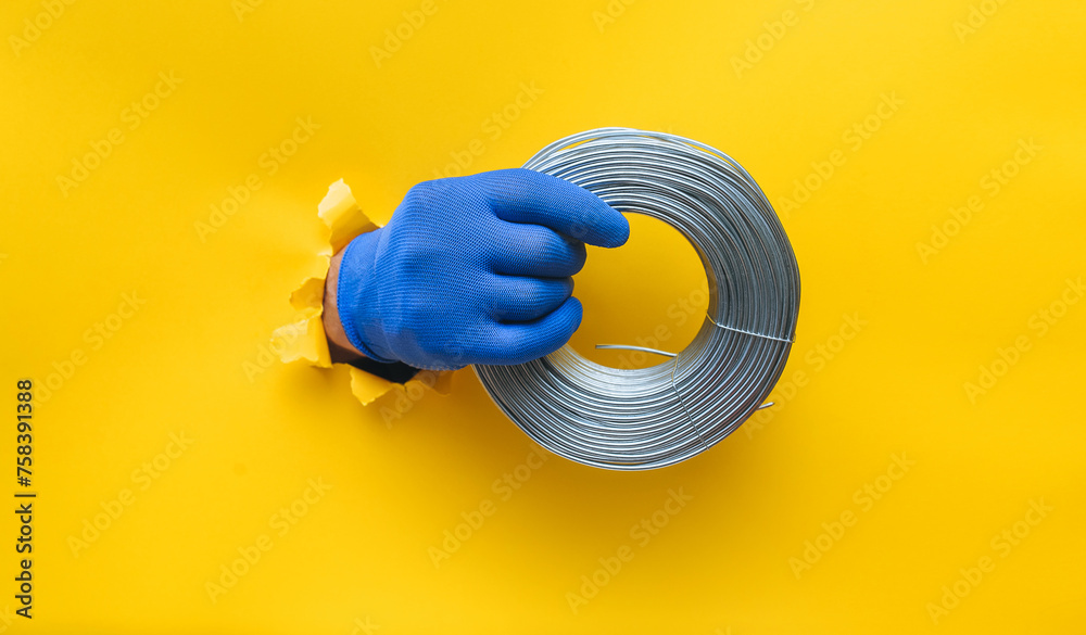 Wall mural A right man's hand in a blue knitted glove holds a coil of steel wire. Torn hole in yellow paper. The concept of a worker, a labor migrant, a master of his craft. Copy space.