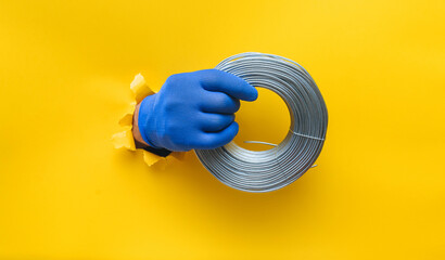 A right man's hand in a blue knitted glove holds a coil of steel wire. Torn hole in yellow paper....