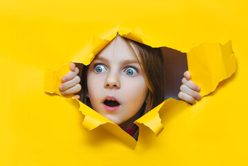 Funny red-haired child girl peeping through hole on yellow paper. The concept of surprise, fear,...
