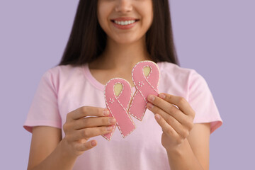 Young woman with cookies in shape of pink ribbons on lilac background, closeup. Breast cancer awareness concept