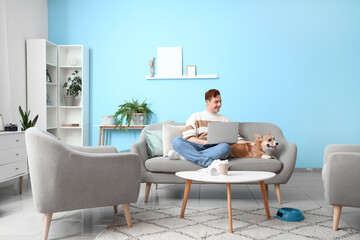 Redhead young happy man with cute Corgi dog and laptop on sofa at home