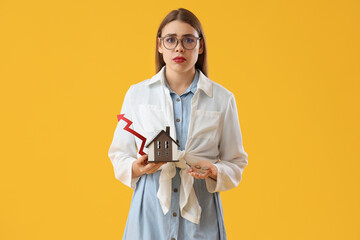 Fototapeta premium Upset young woman with arrow, coins and house model on yellow background. Price rise concept
