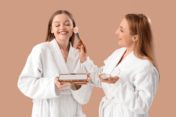 Young woman with her mother in bathrobes doing makeup on beige background