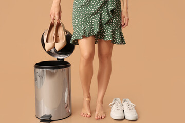 Young woman throwing high-heeled shoes into trash bin on beige background. Uncomfortable shoes...