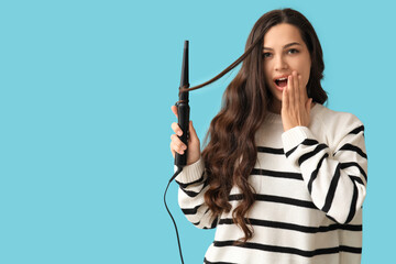 Surprised young woman with curling iron on blue background