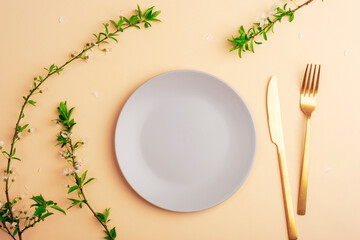 Gray plate and cutlery on light peach background with cherry blossom branches. Top view, flat lay, mockup