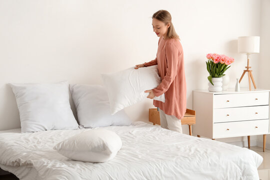 Pretty young woman putting pillow on bed in light bedroom