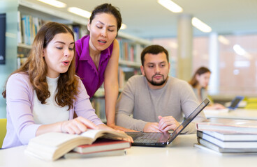 Fototapeta premium Three students are preparing for classes on a laptop in the university library, where girls classmates are looking for ..information in a textbook and discussing something
