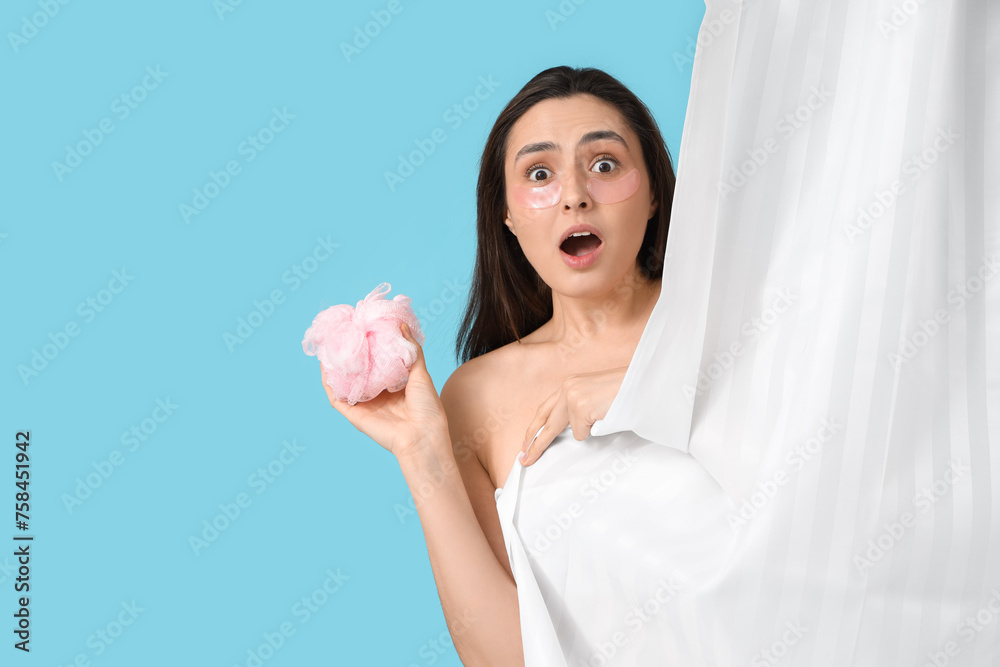 Sticker shocked young woman with loofah behind bathroom curtain on blue background