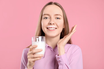 Smiling woman with milk mustache holding glass of tasty dairy drink on pink background