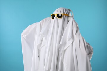 Stylish ghost. Person covered with white sheet in sunglasses and headphones on light blue background