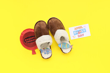 House slippers with paper fishes, whoopee cushion and festive postcard for April Fools Day on yellow background