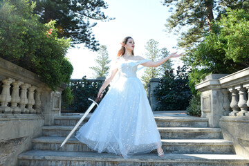Fototapeta premium full length portrait of beautiful female model wearing blue fantasy ballgown, like a fairytale elf princess. Holding. Sword weapon, standing on staircase of a romantic castle balcony location.