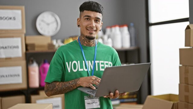 Handsome young latin man, tattooed and donned in t-shirt, confidently smiling as he volunteers with laptop at charity center, serving community with altruism.