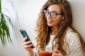 Happy woman holding tea and using smartphone sitting at home. Communication, free time, lifestyle...