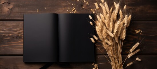 A black book lies on a hardwood table adorned with a bunch of wheat ears. The contrast between the wood and the plant creates a rustic and artistic ambiance