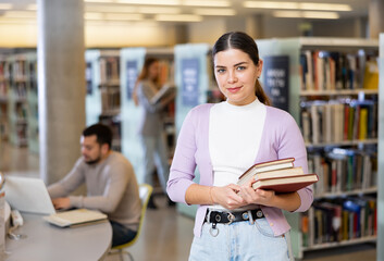 Portrait of a smiling student girl standing in the university library, holding textbooks in her hands
