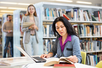 Portrait of young adult woman studying at library using books and laptop computer