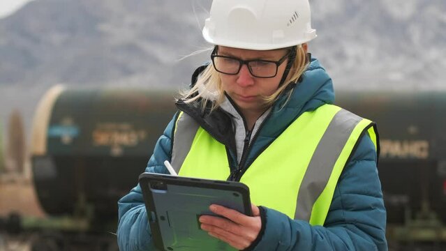 Female worker in white helmet checks shipment, write with a tablet, outdoors, inspecting work, management logistics, standing against backdrop oil wagons.