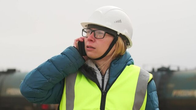 Female quality control inspector talking on smartphone in uniform and hard hat, solves problems, standing on railway station monitors loading and dispatch of freight trains.