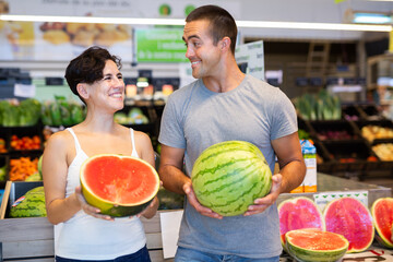 Positive woman and man holding watermelons, standing in aisle of supermarket. Couple making...