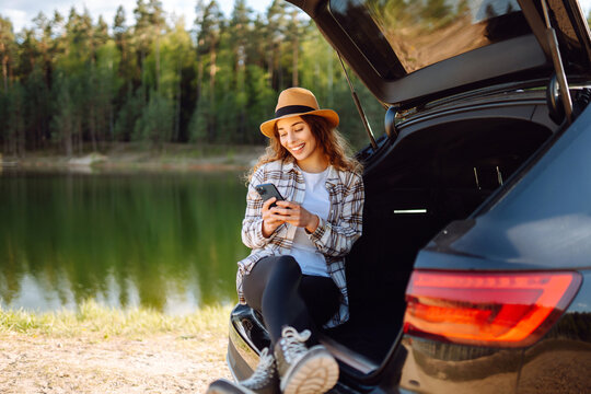 Happy woman  with a phone in her hand sits in the trunk of a car. Concept of technology, adventure, travel by car.