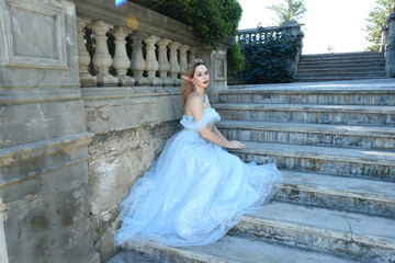 full length portrait of beautiful female model wearing blue fantasy ballgown, like a fairytale elf princess. Seated pose, sitting on staircase of a romantic castle balcony location.