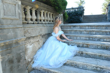 full length portrait of beautiful female model wearing blue fantasy ballgown, like a fairytale elf princess. Seated pose, sitting on staircase of a romantic castle balcony location.