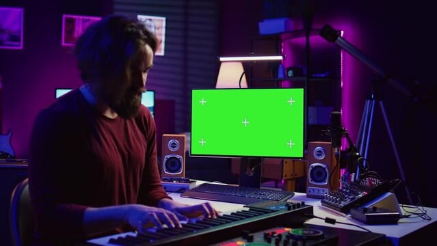Artist learning to play at electronic piano keyboard in home studio, watching tutorial online with greenscreen display. Music producer practices playing on midi controller synthesizer. Camera B.