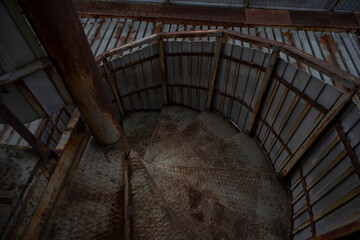 Obraz premium Weathered metal stairs spiral upward inside an industrial structure, hinting at the passage of workers past. The wear and rust speak to the harsh conditions and the relentless passage of time.